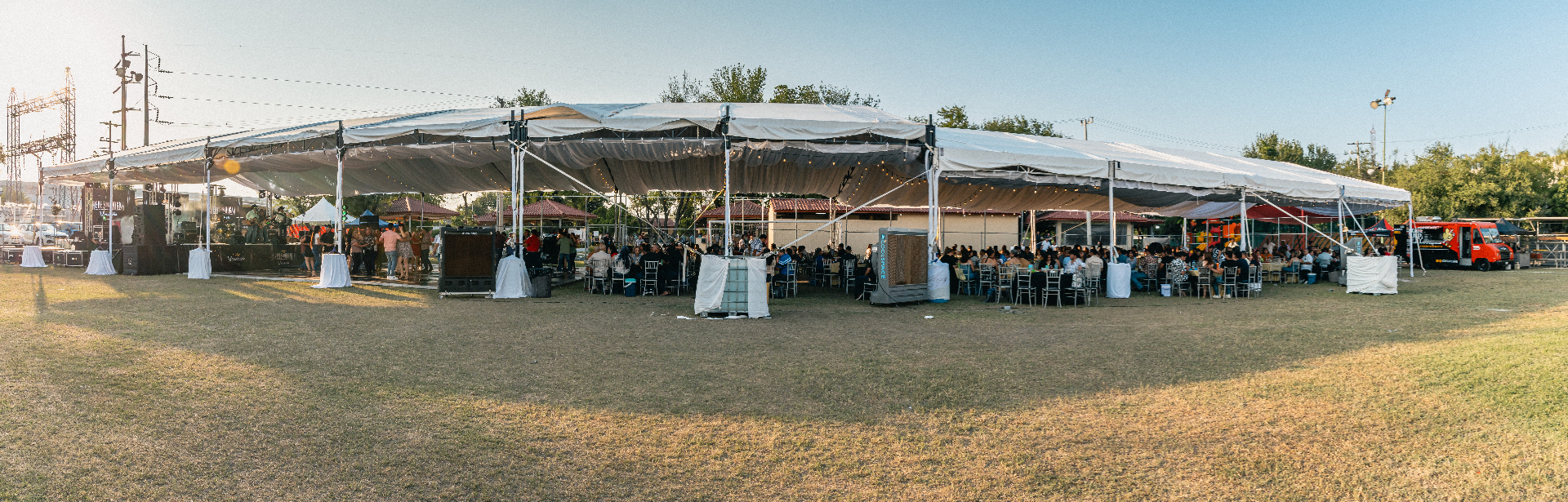 Montaje exterior con carpa, escenario y áreas de comensales al atardecer.