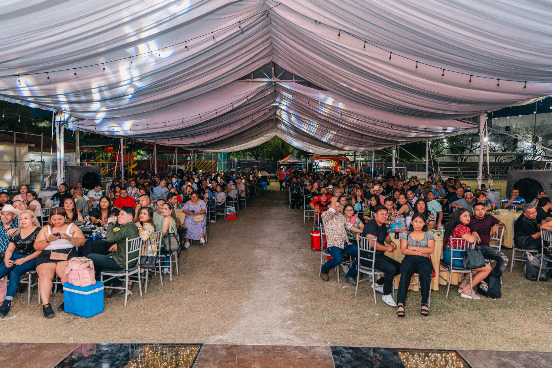 Audiencia sentada en mesas bajo gran carpa iluminada durante la noche.